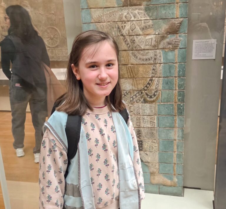 EIFA A young girl with brown hair, wearing a light blue vest and floral-patterned top, stands and smiles in front of an ancient stone relief at a museum. She has a backpack and the exhibit label is visible in the background.