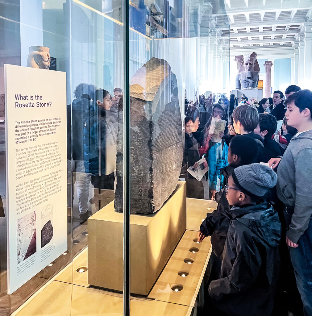 EIFA Des visiteurs se rassemblent autour de la pierre de Rosette exposée dans une vitrine en verre dans un musée. Un grand panneau dinformation sur lartefact est visible sur la gauche, et un groupe de personnes, dont des enfants, observent attentivement lexposition.