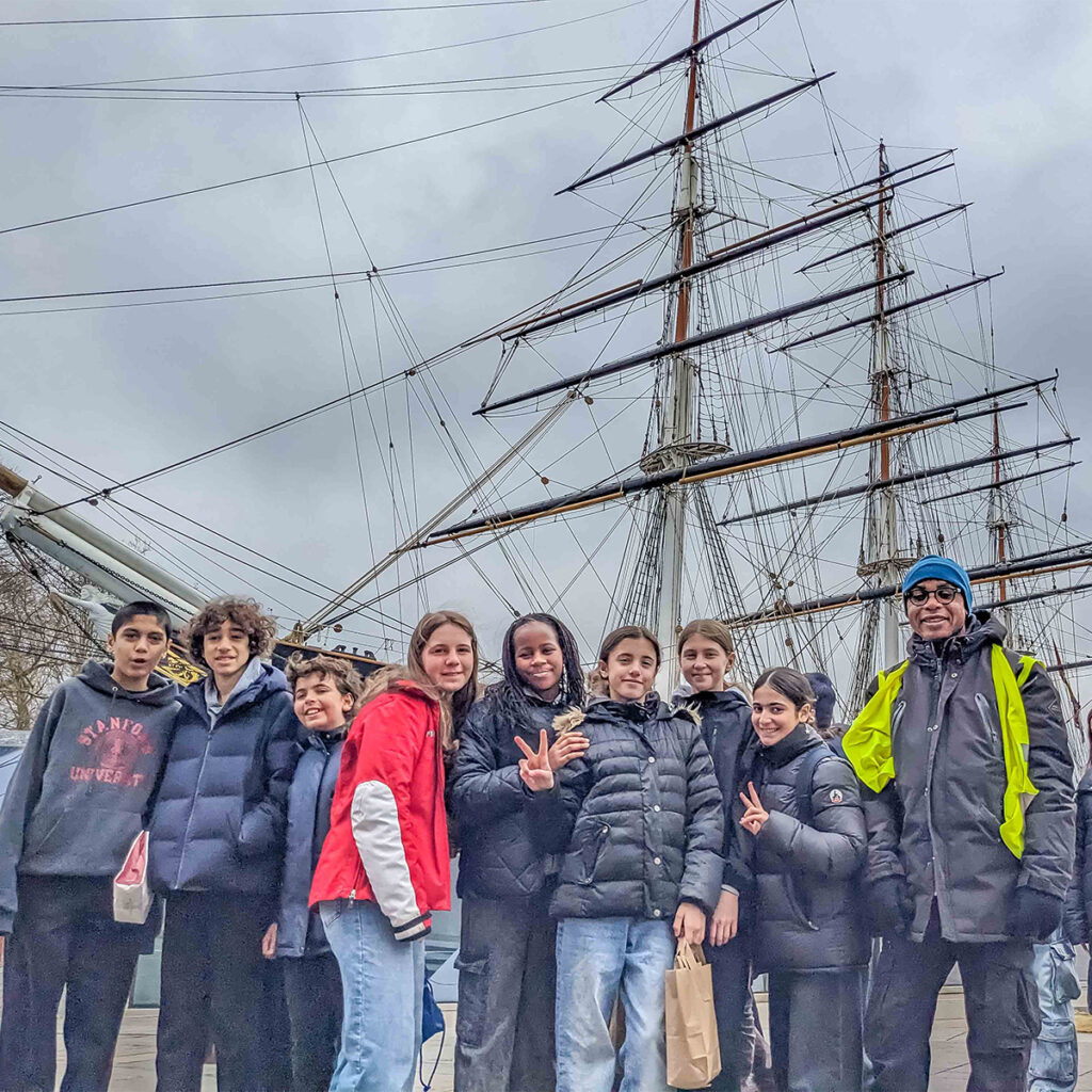 EIFA A group of smiling people, mostly children in winter coats, pose in front of a large sailing ship with tall masts. The sky is overcast, and one person wears a bright yellow jacket and blue cap.