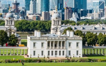 EIFA Une vue panoramique de Greenwich, rappelant À la découverte du Soho français, met en valeur un bâtiment historique blanc avec des colonnes au premier plan. Des pelouses et des arbres verdoyants contrastent avec les gratte-ciel modernes s'élevant sous un ciel bleu éclatant.