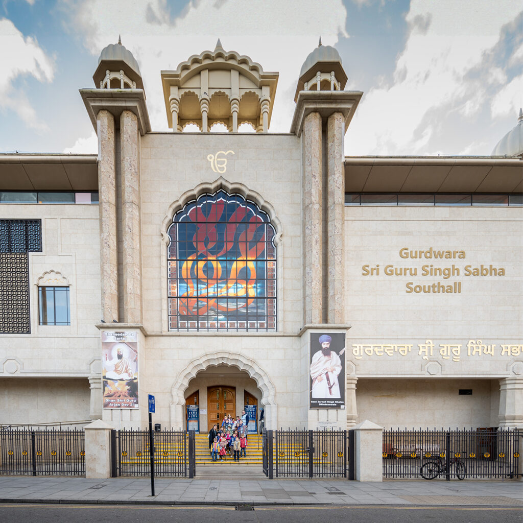 EIFA The image captures the exterior of Gurdwara Sri Guru Singh Sabha in Southall, featuring a large arched entrance, stained glass artwork, and people entering the building. Reminiscent of a school for spiritual learning, its name gleams in bilingual gold letters on the facade.