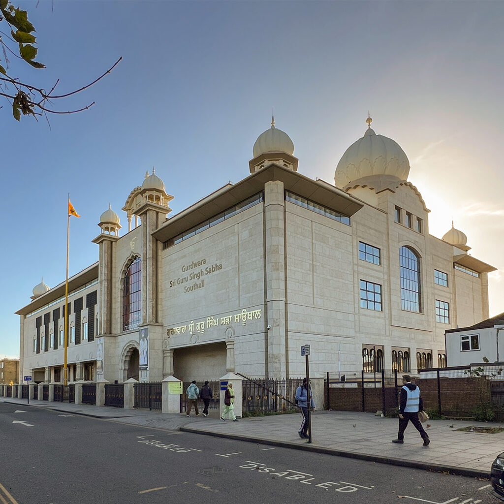 EIFA A large Sikh temple, Gurdwara Sri Guru Singh Sabha, stands proudly in Southall, London. Its cream facade with arched windows and multiple domes gleams as the sun sets, casting a warm glow. Nearby, a few bilingual visitors stroll by while the Indian flag flutters gracefully in the background.