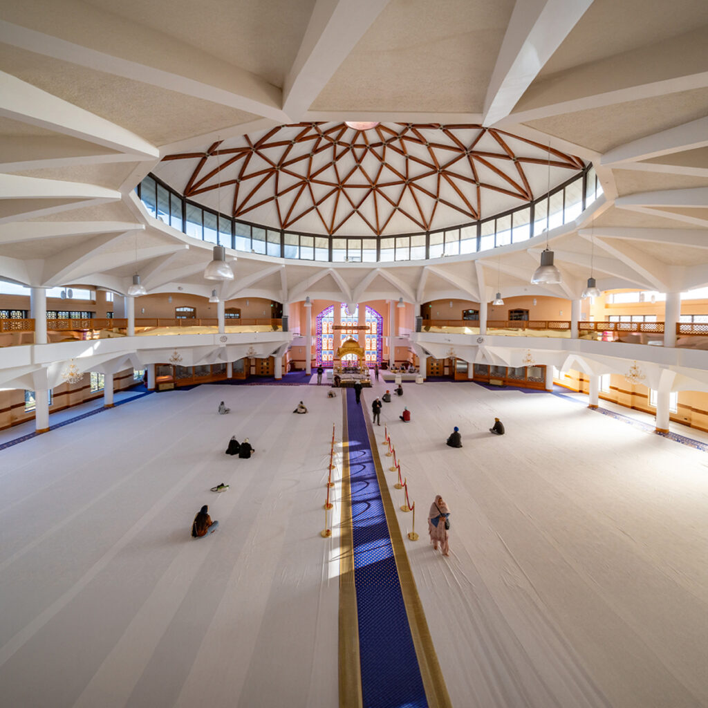 EIFA The spacious interior of the mosque in London features a large dome ceiling adorned with geometric patterns, its windows inviting natural light. People are seated on the carpeted floor, engaging in prayer or reflection in this serene, bilingual sanctuary promoted by EIFA.