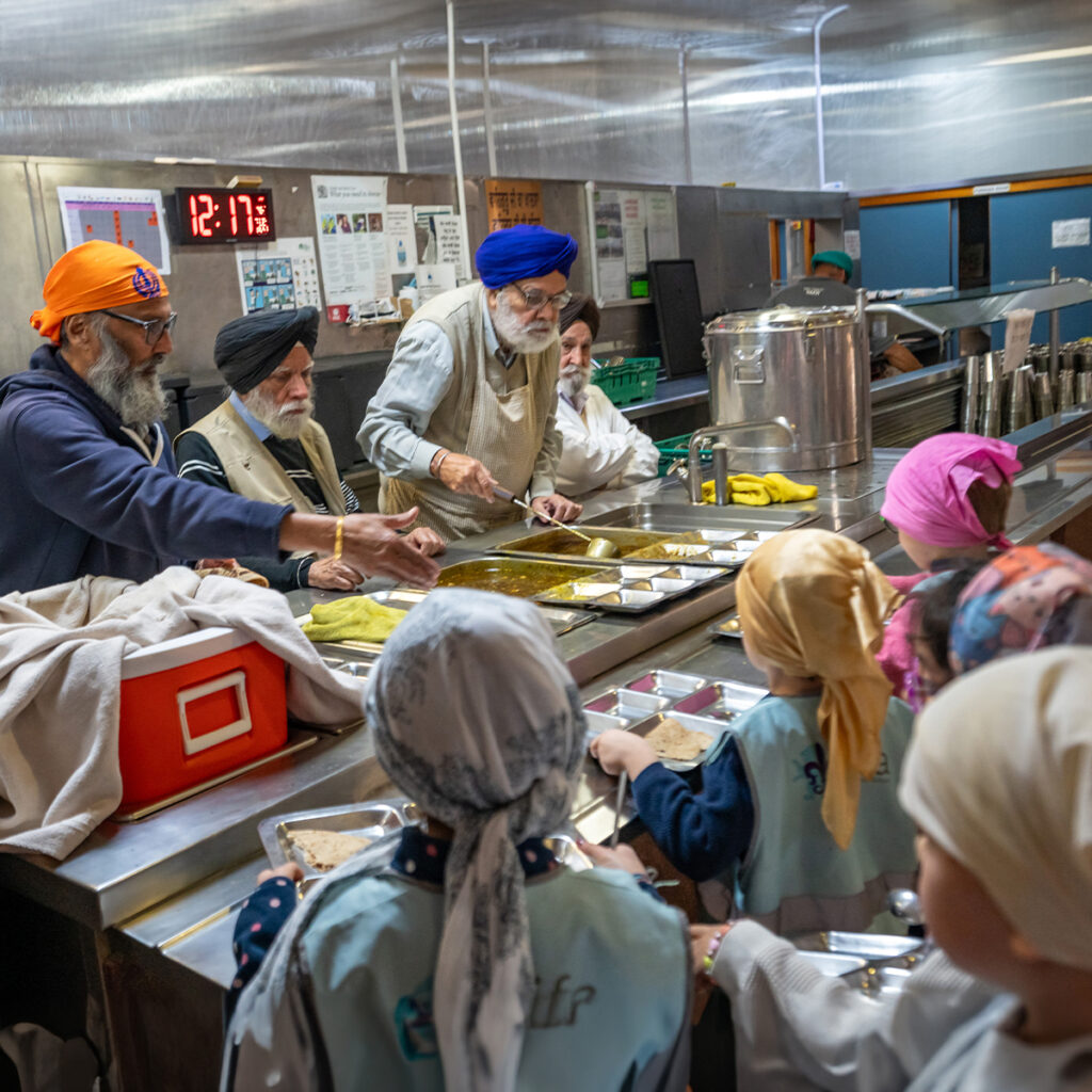 EIFA Sikh men, wearing turbans and aprons, serve food to children at a bilingual community kitchen in London. Behind a stainless-steel counter with large pots and trays, they work efficiently. The children, wearing head coverings, wait patiently in line as the clock reads 12:17.