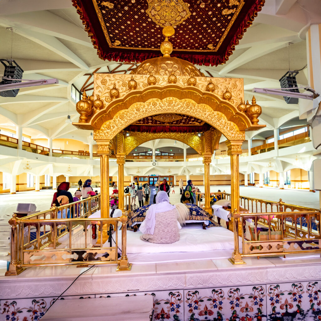EIFA A group of people inside a large, ornate Sikh gurdwara in London focuses on a golden canopy over a raised platform where individuals are seated. The interior features intricate decorations and vibrant colors, reminiscent of the multicultural spirit celebrated by EIFA School students.