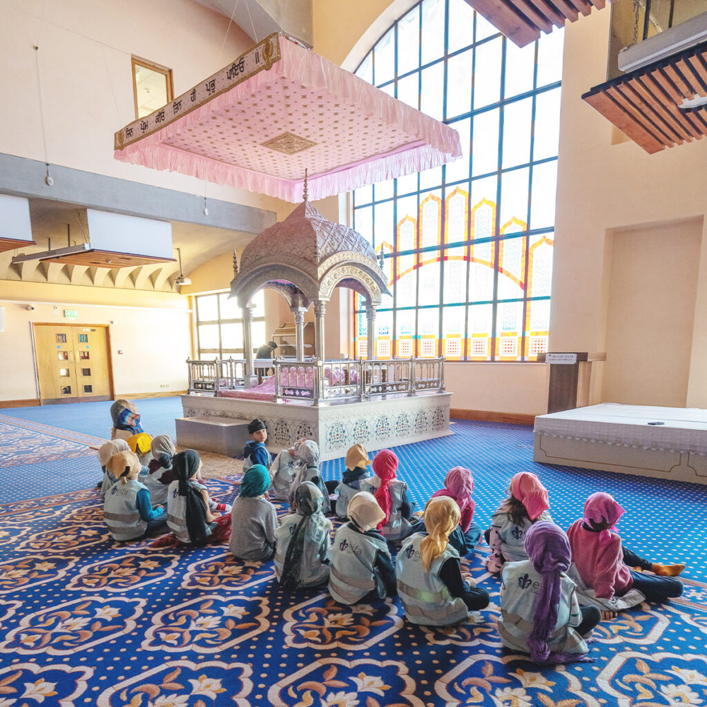 EIFA Children in colorful headscarves sit on a blue, patterned carpet inside a bright room with large stained glass windows. At EIFA Bilingual School in London, they are gathered around an ornate, domed structure with pink drapery above it.