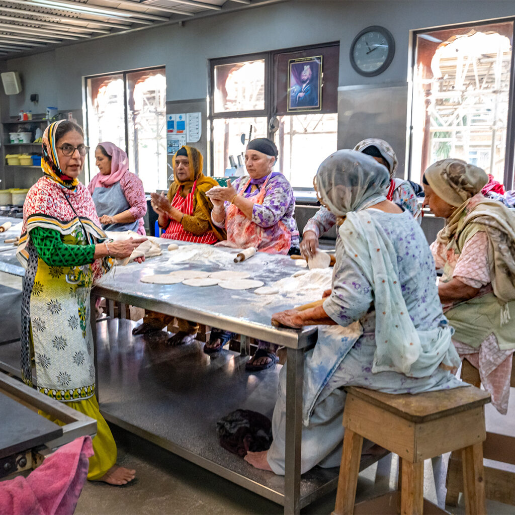 EIFA A group of women in traditional clothing prepares flatbreads around a large table in a well-lit, bilingual kitchen. Some are seated while others stand, working with dough and flour. A framed picture and clock hang on the wall behind them.