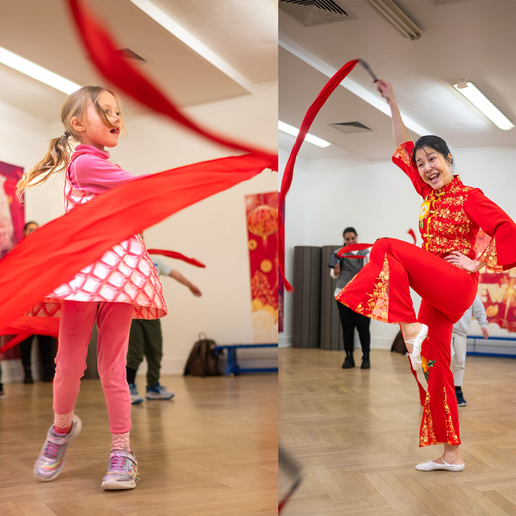 EIFA Une femme et un enfant pratiquent une danse traditionnelle avec des rubans rouges dans une pièce très éclairée. La femme porte une tenue rouge et or, et lenfant est habillé en rose. Dautres personnes et des décorations festives sont visibles en arrière-plan.