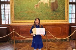 EIFA A young girl in a blue dress stands on a wooden floor, holding a certificate and medal at the International Open Day. She is smiling in front of a large painting of a woman feeding sheep, framed by wood-paneled walls and velvet ropes.
