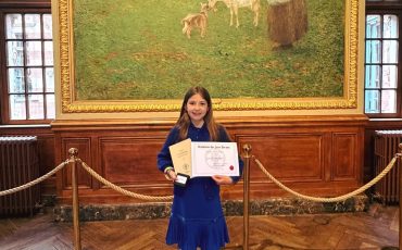 EIFA A young girl in a blue dress stands on a wooden floor, holding a certificate and medal at the International Open Day. She is smiling in front of a large painting of a woman feeding sheep, framed by wood-paneled walls and velvet ropes.