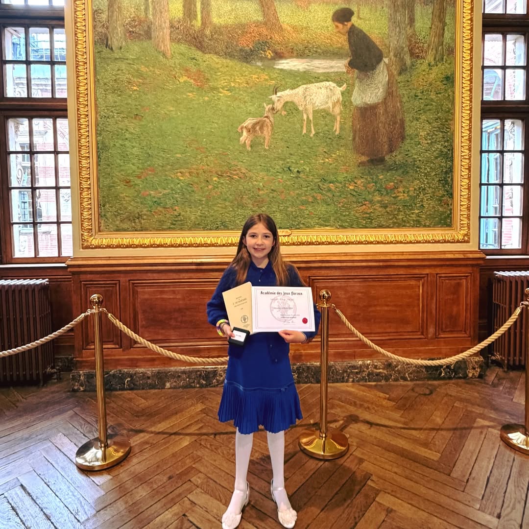 EIFA A young girl in a blue dress stands on a wooden floor, holding a certificate and medal at the International Open Day. She is smiling in front of a large painting of a woman feeding sheep, framed by wood-paneled walls and velvet ropes.