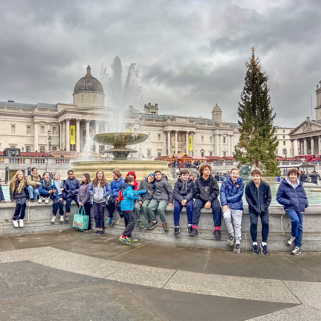EIFA A group of children sit on the edge of a fountain in front of a large, domed building in London. A Christmas tree is nearby under the overcast sky. The bilingual children from EIFA appear relaxed and enjoying themselves.