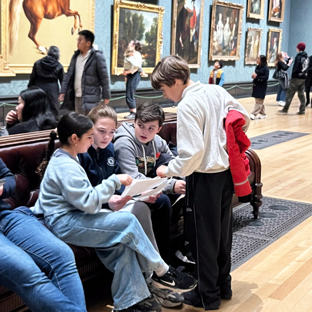 EIFA Three children sit on a bench in the art museum, engaging with a bilingual pamphlet. One child stands, leaning slightly as they look on. Paintings adorn the walls, and other visitors are seen in the background, reminiscent of a day at London’s own EIFA gallery.