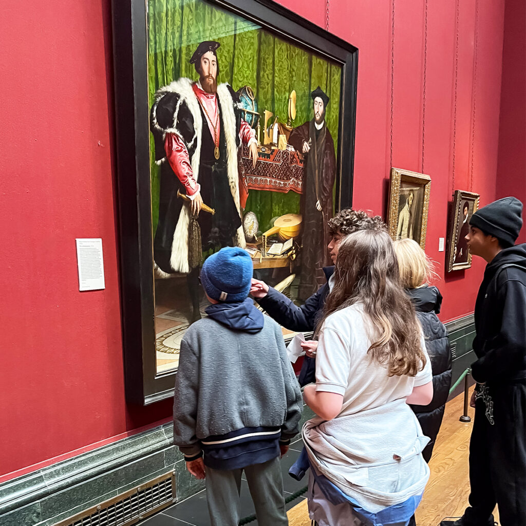 EIFA A bilingual group, perhaps from a London school, including a boy pointing, stands before a large painting of two men with musical instruments and globes. The red-walled corridor hosts smaller artworks.