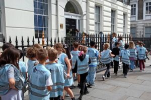 EIFA A group of children wearing matching blue vests stand in line on a sidewalk outside a white, multi-story building with black railings, eagerly awaiting the start of International Open Day.