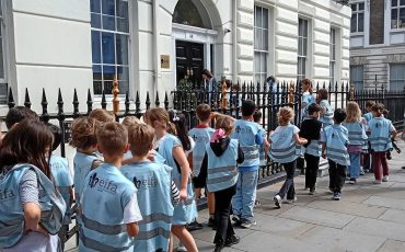 EIFA A group of children wearing matching blue vests stand in line on a sidewalk outside a white, multi-story building with black railings, eagerly awaiting the start of International Open Day.