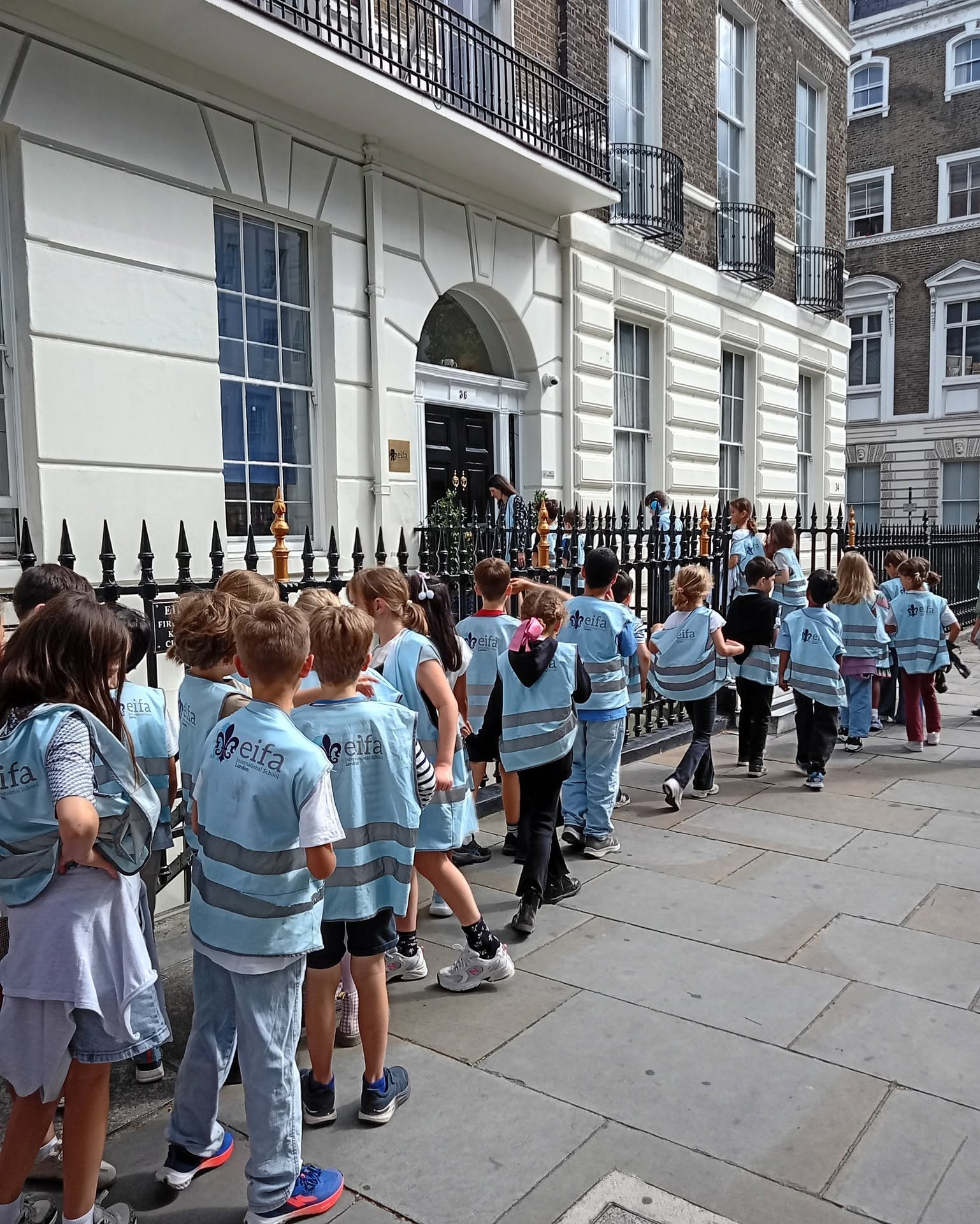 EIFA A group of children wearing matching blue vests stand in line on a sidewalk outside a white, multi-story building with black railings, eagerly awaiting the start of International Open Day.