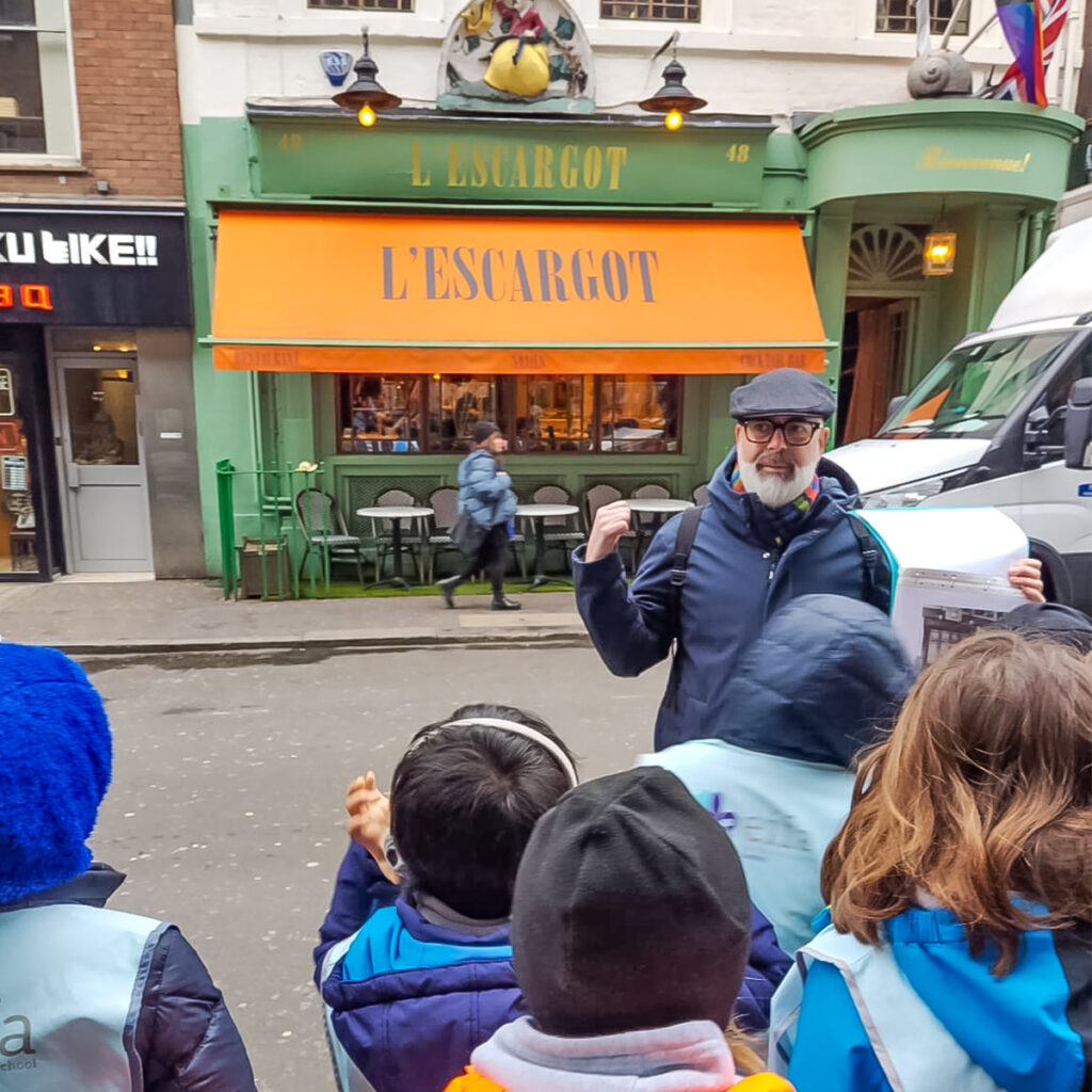 EIFA Un homme portant une casquette et des lunettes fait des gestes enthousiastes avec ses bras, debout devant le restaurant LEscargot. Un groupe denfants écoute attentivement. Le restaurant a un auvent orange vif et des accents verts.