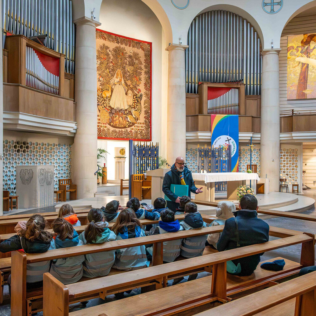 EIFA Une personne parle à un groupe denfants assis dans une église. Lintérieur est orné de grands tuyaux dorgue, dune tapisserie colorée et dœuvres dart religieuses. Léglise est éclairée par la lumière naturelle provenant de hautes fenêtres. Les enfants portent des vestes assorties.