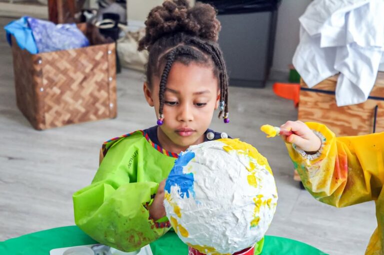 EIFA A young child wearing a green smock paints a large paper mache globe with blue and yellow paint, joined by another child’s hand. The children are seated at a table indoors during International Open Day.