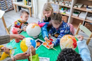 EIFA Quatre jeunes enfants sont assis autour d’une table et peignent de grands globes en papier mâché pour la Journée mondiale de l’alimentation. Concentrés sur leur art, ils utilisent des pinceaux et des peintures colorées sous l’assistance d’un adulte. Des fournitures artistiques et des bouteilles de peinture remplissent la table. EIFA Quatre jeunes enfants sont assis autour d'une table et peignent de grands globes en papier mâché pour la Journée mondiale de l'alimentation. Concentrés sur leur art, ils utilisent des pinceaux et des peintures colorées sous l'assistance d'un adulte. Des fournitures artistiques et des bouteilles de peinture remplissent la table.