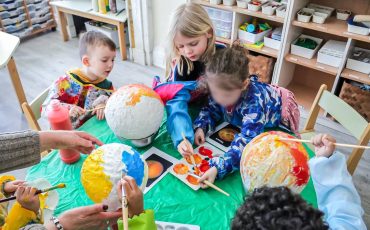 EIFA Several young children sit around a table painting papier-mâché globes with bright colors during International Open Day. Art supplies and paint bottles are on the table, and an adult helps guide the activity in a colorful classroom setting.