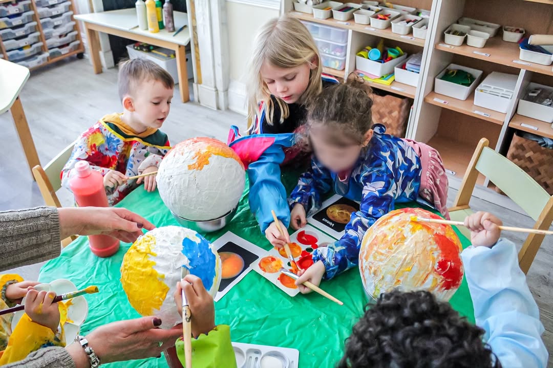 EIFA Several young children sit around a table painting papier-mâché globes with bright colors during International Open Day. Art supplies and paint bottles are on the table, and an adult helps guide the activity in a colorful classroom setting.