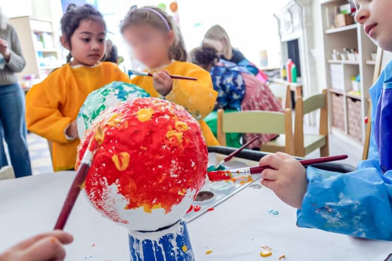 EIFA During International Open Day, young children in colorful smocks paint a large paper mache ball with red, yellow, and green paint at a classroom table, engaging in a creative group art activity.
