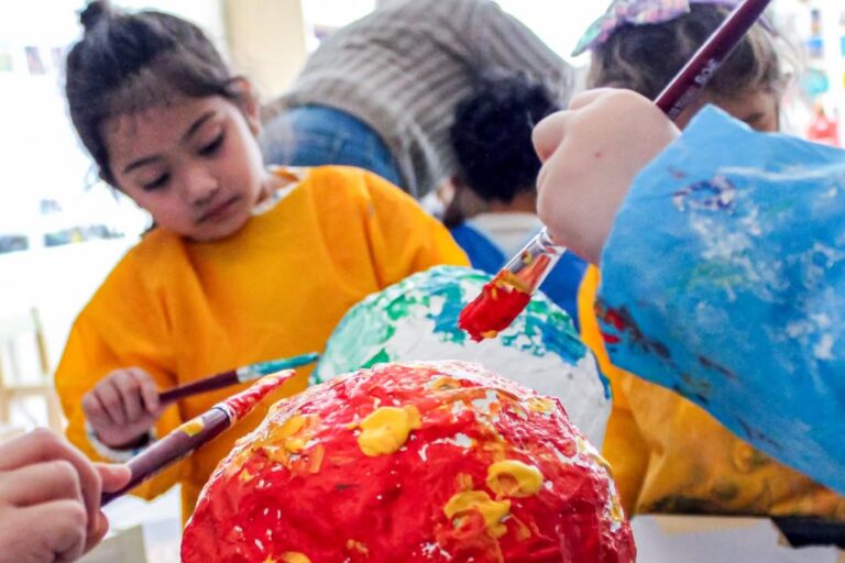 EIFA Children in colorful smocks paint papier-mâché globes at a table during International Open Day, focusing on their creative activity. One child paints a red globe with yellow, while others work with different colors in the background.