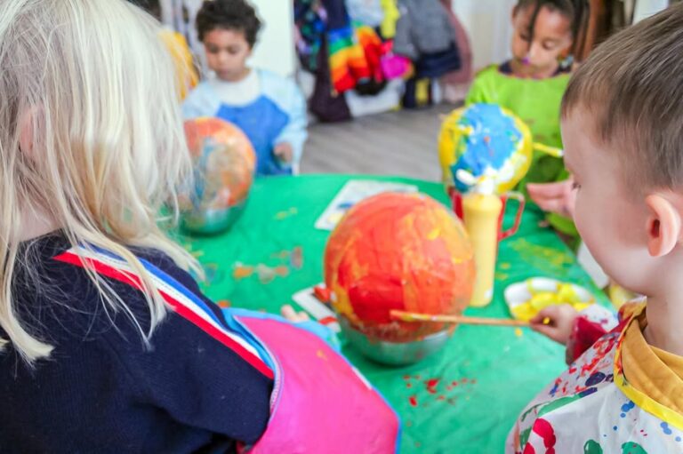 EIFA Several young children sit around a green table, painting colorful papier-mâché balloons during International Open Day. Art supplies and paint bottles are scattered on the table, and the children wear aprons to protect their clothes.