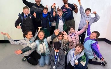 EIFA A group of children wearing medals pose cheerfully with two adults against a plain wall, celebrating International Open Day. The kids smile, raise their arms, and sit or kneel in front while others and the adults stand behind them in high spirits.