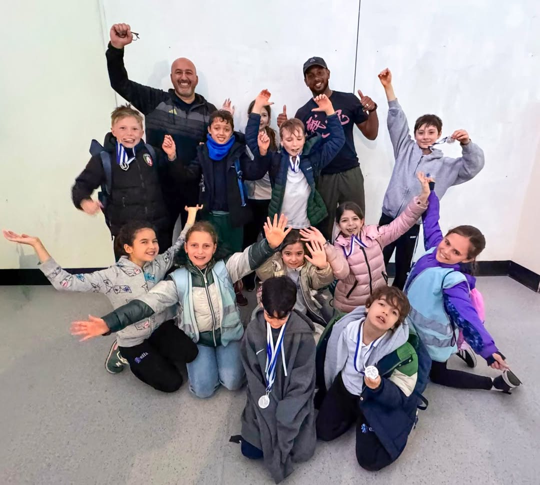 EIFA A group of children wearing medals pose cheerfully with two adults against a plain wall, celebrating International Open Day. The kids smile, raise their arms, and sit or kneel in front while others and the adults stand behind them in high spirits.