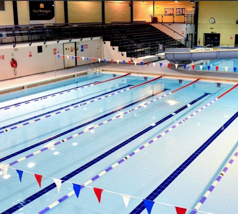 EIFA Indoor swimming pool with clear water, six swim lanes marked with blue lines, and red, blue, and white pennant flags overhead. Empty spectator seating and a large clock hint at preparation for International Open Day events.