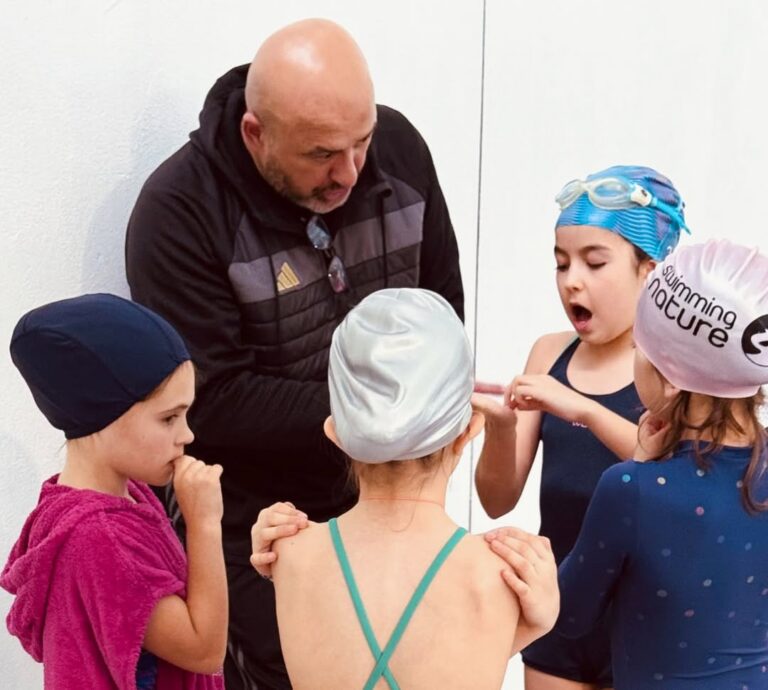 EIFA A swim coach talks to four young swimmers in caps and gear, gathered in a circle by a white wall, listening attentively. The scene captures the excitement of International Open Day as one child gestures while others watch and listen.