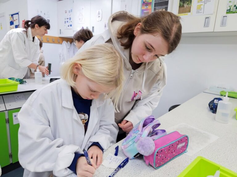 EIFA Two children in lab coats work together at a science lab table during International Open Day. One writes notes while the other watches closely, surrounded by lab equipment and trays, as other students experiment in the background.