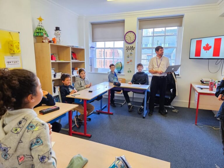 EIFA A teacher stands at the front of a classroom holding a laptop during International Open Day, with several children seated at desks facing him. A screen displays the Canadian flag, and the room has large windows and educational decorations.