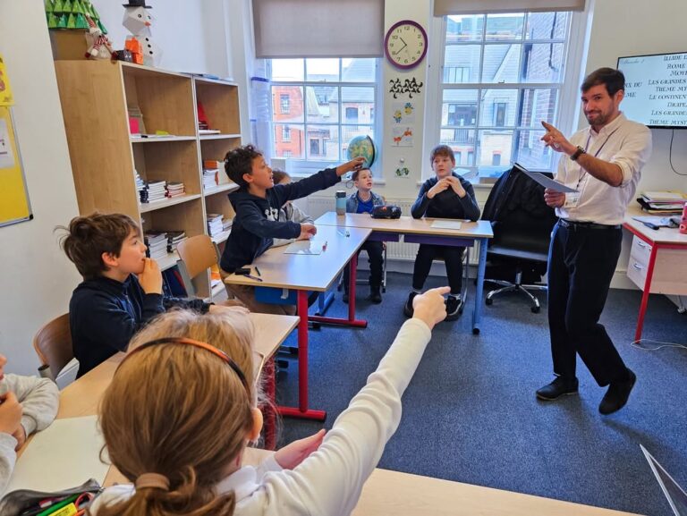 EIFA A teacher stands at the front of a classroom engaging with students, some raising their hands and pointing, during International Open Day. The room features desks, shelves filled with books, and large windows overlooking nearby buildings.