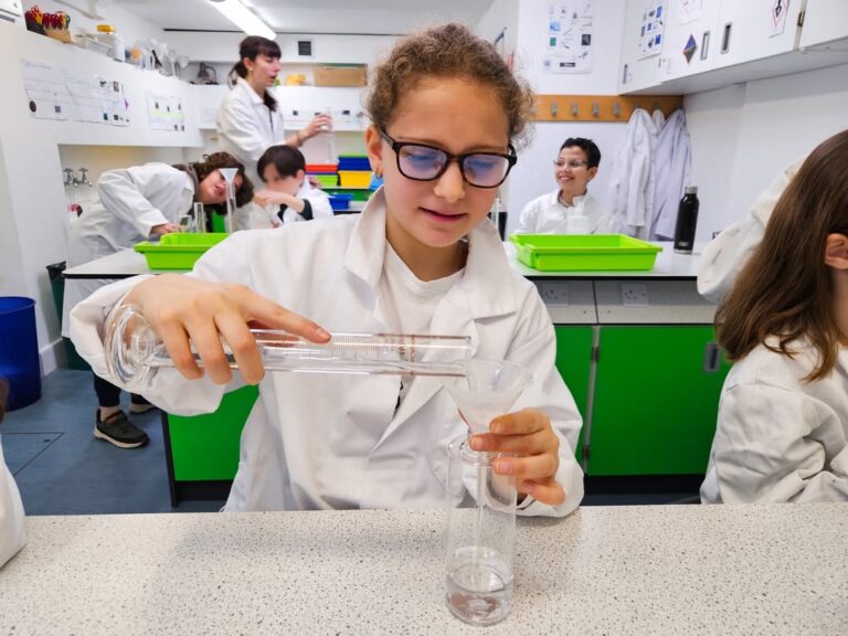 EIFA During International Open Day, a girl in a white lab coat and glasses pours liquid from a beaker into a graduated cylinder in a science classroom, while other children in lab coats work at tables in the background.