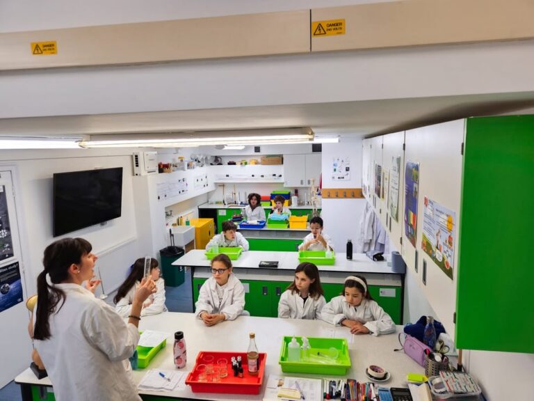 EIFA A teacher in a white lab coat stands at the front of a brightly lit science classroom, demonstrating an experiment for International Open Day to students seated at green tables, all wearing lab coats and watching attentively.