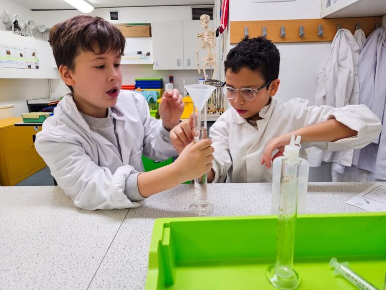 EIFA Two boys in white lab coats work together in a science lab during International Open Day, pouring liquid through a funnel into a graduated cylinder. Lab equipment, a green tray, and a skeleton model are visible in the background.