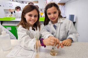 EIFA Deux jeunes filles en blouse blanche sourient en réalisant une expérience scientifique sur la Journée mondiale de l’alimentation à une table de laboratoire, à l’aide de béchers et d’éprouvettes. Du matériel de laboratoire et d’autres étudiants sont visibles à l’arrière-plan. EIFA Deux jeunes filles en blouse blanche sourient en réalisant une expérience scientifique sur la Journée mondiale de l'alimentation à une table de laboratoire, à l'aide de béchers et d'éprouvettes. Du matériel de laboratoire et d'autres étudiants sont visibles à l'arrière-plan.