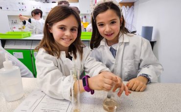EIFA Two young girls in white lab coats smile while conducting an experiment during International Open Day. One stirs a liquid as the other watches, surrounded by lab equipment and fellow students in a vibrant science classroom.