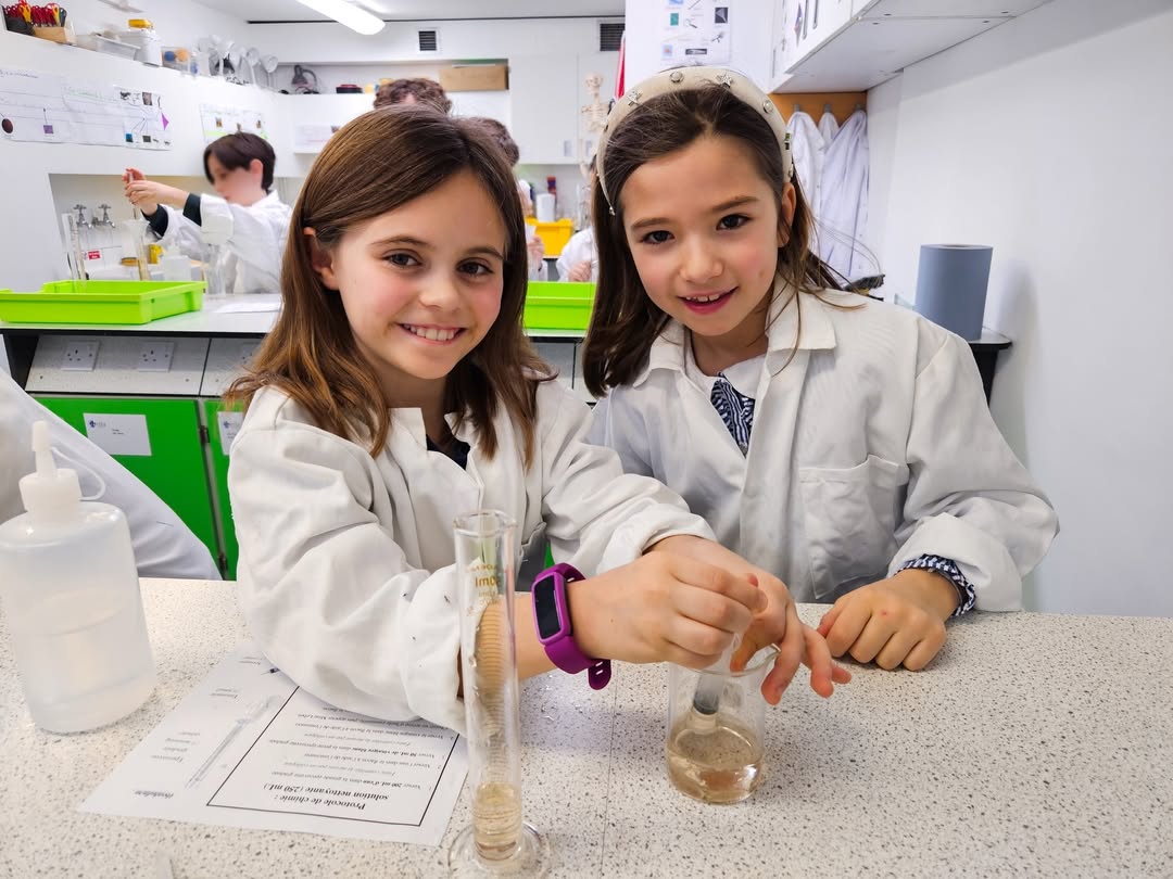 EIFA Two young girls in white lab coats smile while conducting an experiment during International Open Day. One stirs a liquid as the other watches, surrounded by lab equipment and fellow students in a vibrant science classroom.