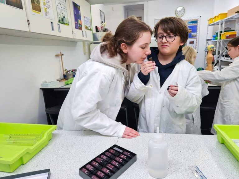 EIFA Two students in white lab coats stand at a lab table during International Open Day; one smells a vial held by the other. Science supplies, such as test tubes and a plastic bottle, sit on the table while other students work in the background.