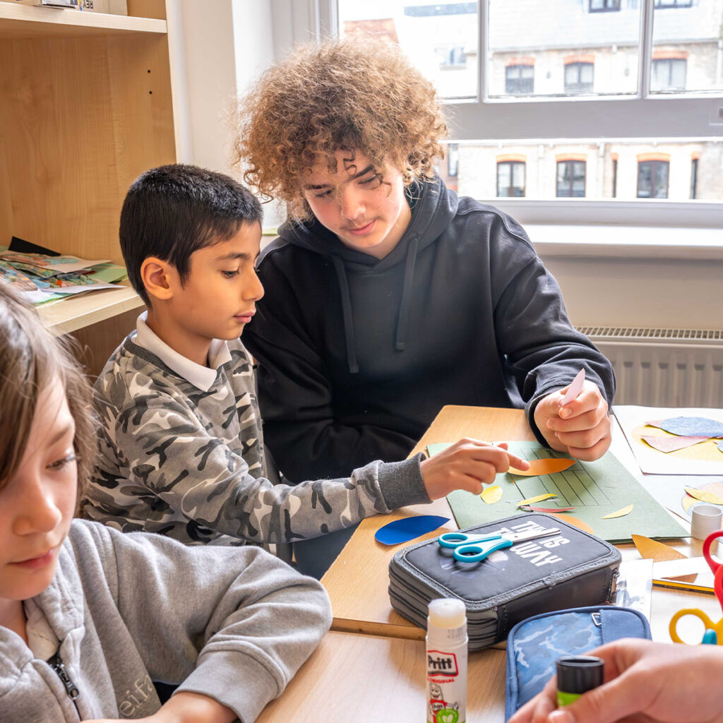 EIFA Two children sit at a table crafting with colorful paper, glue, and scissors. One child with curly hair assists a younger child wearing a camo hoodie. They are focused on the activity in a bright room with large windows.