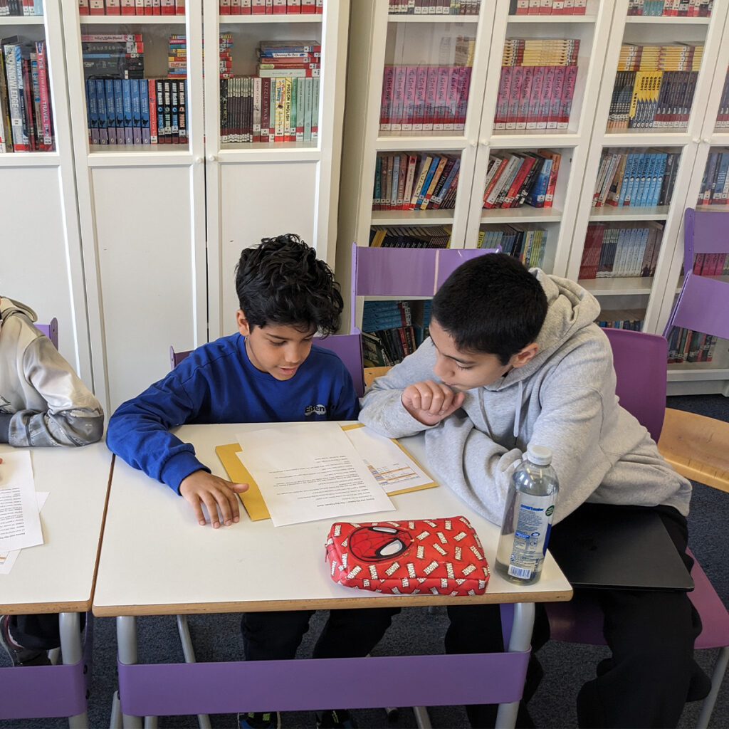 EIFA Two students are sitting at a table in a library, focused on reading a document together. One wears a blue shirt and points at the page, while the other in a grey hoodie leans in. Bookshelves filled with books are in the background.
