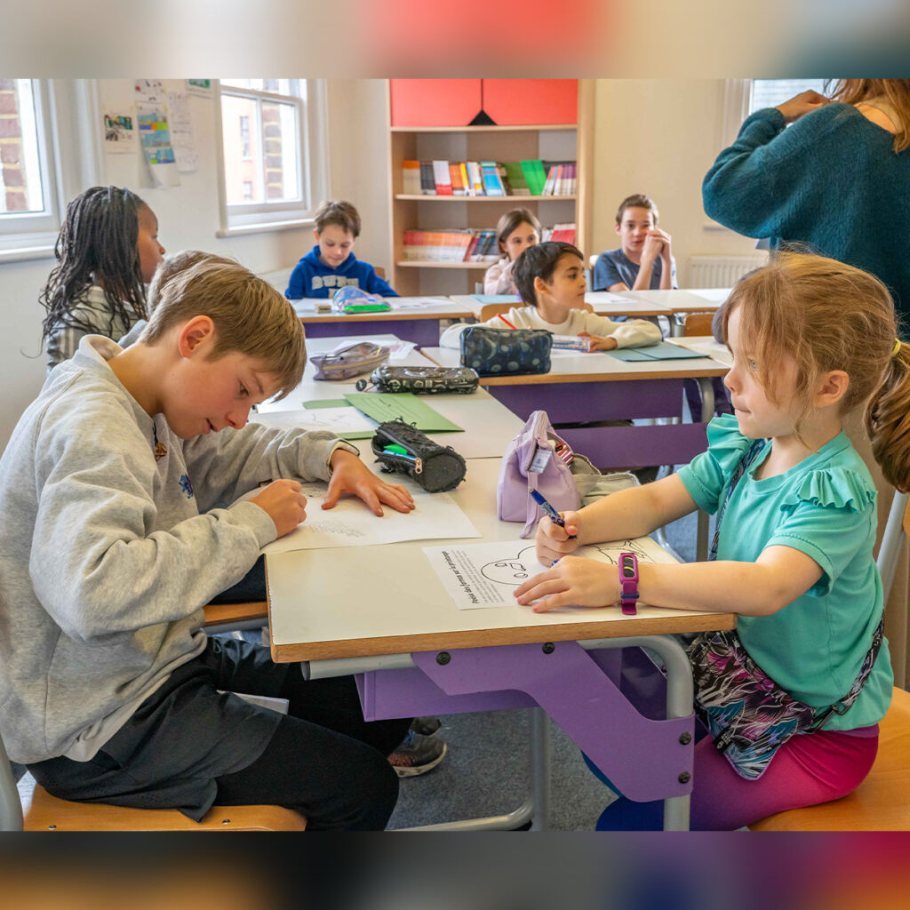 EIFA Children sitting at desks in a classroom, drawing and writing on papers. A teacher is standing near a bookshelf, assisting students. The room is bright with natural light coming through the windows.