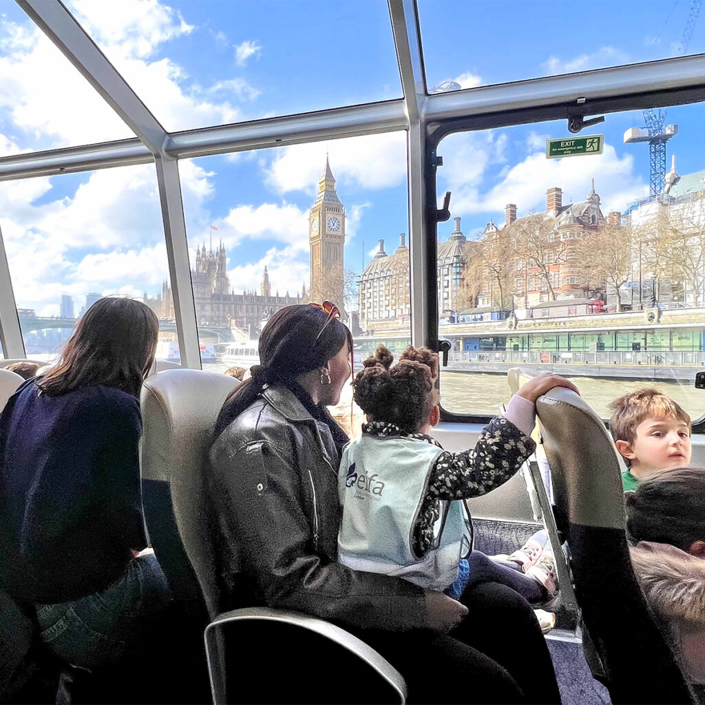 EIFA A group of people, including children, sit inside a glass-roofed boat looking at the view of Big Ben and the Houses of Parliament on a sunny day. The River Thames and a partly cloudy sky are visible outside.