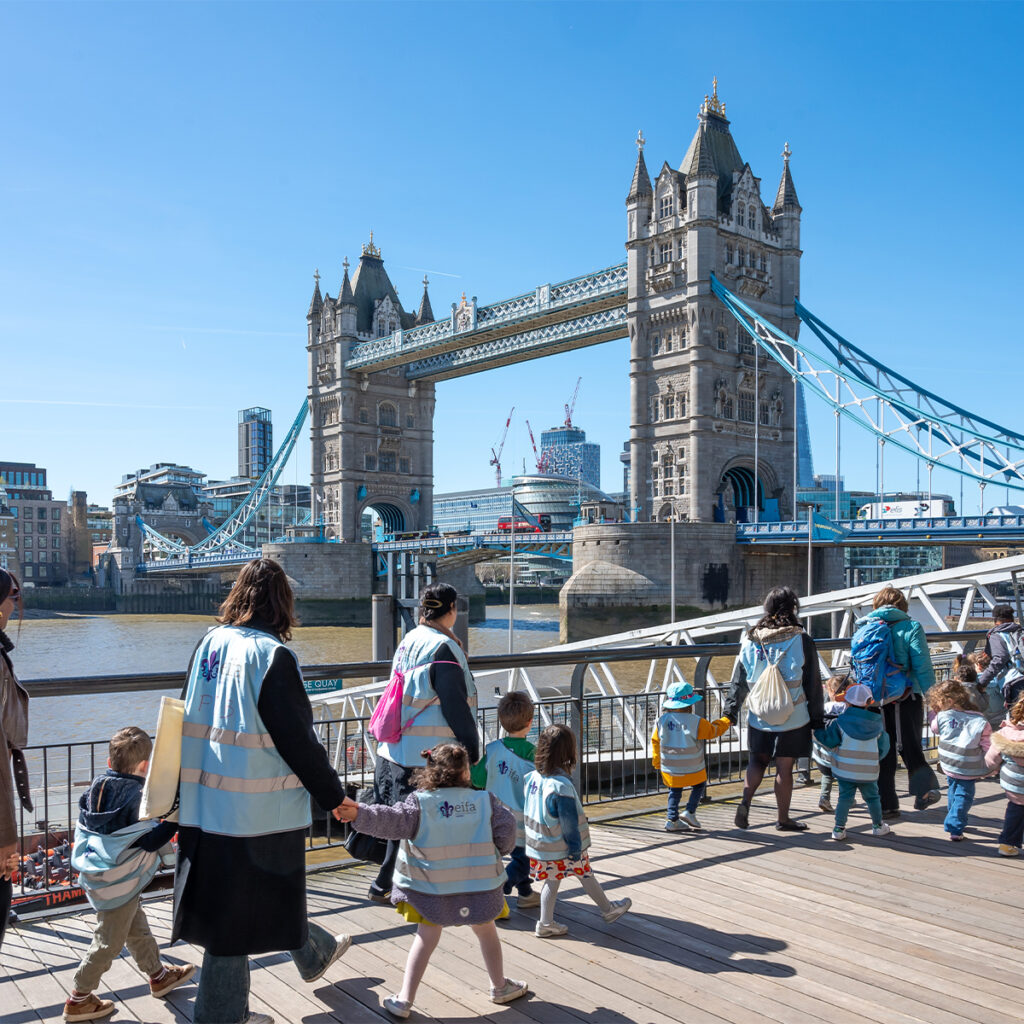 EIFA A group of adults and children wearing reflective vests walk along a riverside boardwalk near Tower Bridge in London. The iconic bridge is visible in the background under a clear blue sky.