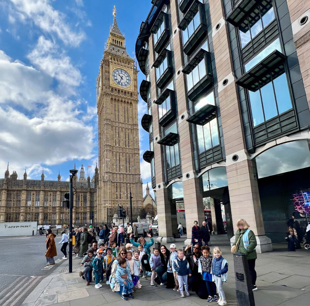 EIFA A group of people are gathered in front of a modern building with the iconic Big Ben and Westminster Palace in the background. The sky is partly cloudy, and the scene is lively with pedestrians.